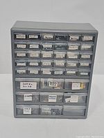 Front view of gray plastic multi-drawer hardware organizer with handwritten labels on each drawer describing contents such as drywall nails, hinges, dowel guides, and shelf pins.