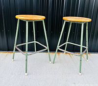 Pair of stools with round oak seats and green tubular steel legs with footrests, positioned against black corrugated metal background.