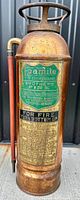 Front view of the antique copper fire extinguisher showing green and gold label with instructions and brand.