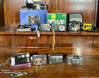 Wide view of multiple cameras arranged on wooden shelving, shows various digital and film cameras along with a Sony Discman CD player.