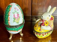 Two ceramic trinket boxes side by side: egg-shaped floral design with metal hinge and clasp on feet, and a rabbit in yellow outfit holding a red object with metal hinge clasp.