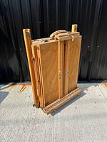 Easel standing upright showing front wood panels and leather handle, stored in folded position.
