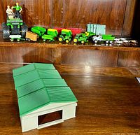 Photo showing all the John Deere farm toys and accessories arranged on a wooden shelf, including farm vehicles, trailers, livestock miniature figures, two people figurines, and a large plastic barn building with green roof and white siding.