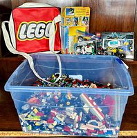 Photo showing large clear plastic storage bin filled with assorted loose LEGO bricks and parts, red LEGO branded bag with shoulder strap, opened LEGO Creator 3-in-1 kit boxes, and various LEGO components including wheels and vehicle pieces.