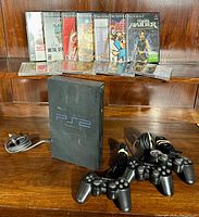 Photo of PlayStation 2 console standing vertically, power cord, four wired controllers laid out on a wooden surface, and a row of game cases lined up behind the console.
