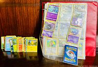 Image showing loose Pokémon cards in a stack and an open red album with multiple Pokémon cards visible in plastic sleeves.