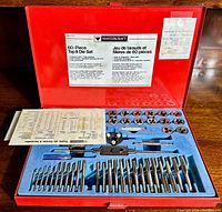 Open red metal case showing blue foam insert with rows of metal taps and dies, tap wrench, and additional tools included in the set. Instruction sheet visible.