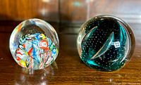 Two vintage art glass paperweights side by side on wooden surface showing millefiori and greenish-blue designs.