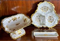 Three white porcelain serving dishes with detailed gold floral decoration displayed on a wood surface, including a three-compartment dish, pedestal dish, and long narrow tray.