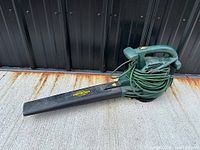 Photo of green Yardworks electric leaf blower showing full device with attached power cord wound on it, black approx. 24-inch blower tube, taken on concrete surface.