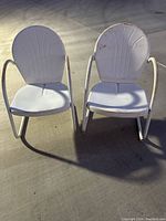 Front view of two white metal vintage clamshell chairs showing rounded backs and tubular armrests with visible some rust and wear.