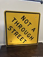 Front view of a square yellow metal street sign with black text 'NOT A THROUGH STREET' and black border. Mounted upright against a white wall.