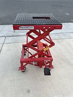 Front side view of red motorcycle jack stand on concrete with four caster wheels, foot pedal, and textured platform