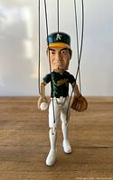 Front view of the Eric Chavez marionette puppet showing the face, green cap and Oakland Athletics jersey with baseball in hand and white pants.