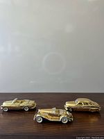 Front view of three vintage gold-tone collectible model cars on wooden surface, showing convertible, luxury car, and sedan models.
