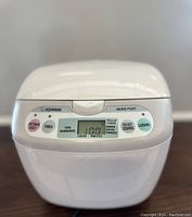 Front view of white Zojirushi Neuro Fuzzy rice cooker with digital display showing 1:00 time remaining and control buttons on front panel.