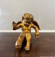 Front view of a golden gilded cherub figurine seated with one leg raised and right hand raised.