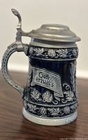 Side view of beer stein showing inscription 'Gott erhalts' with detailed hop and leaf relief decorations on dark blue ceramic body and pewter lid with thumb lever.