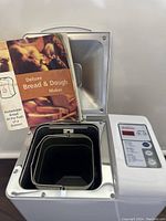 Open bread maker showing stainless steel cooking chamber, bread pan with kneading blade, and included user manual.