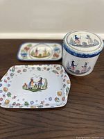 Three vintage French Quimper ceramics shown on wooden surface including two plates and one jar with lid. Hand-painted traditional Breton folk designs and floral motifs visible.
