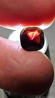 Close-up photo of the 4.12ct cushion cut red spinel held between fingers, showing deep red color with purple undertones under direct light and visible inclusions.