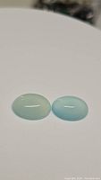 Two bright Arctic teal blue oval cabochon chalcedony gemstones on white background, showing size and color.