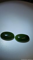 Two oval cabochon deep green jade stones placed side by side, showing the smooth glossy surface and rich color.