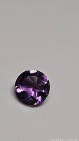 Close-up photo of a single faceted round deep purple amethyst gemstone with diamond cut facets, showing reflective surfaces and internal clarity.