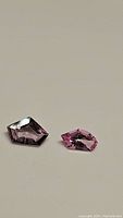 Two natural fancy cut spinel gemstones on plain light background. One is purple shield cut, the other is pink fancy cut.