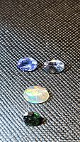 Four natural gemstones displayed on textured dark surface under light: blue tanzanite, light blue sapphire, iridescent Ethiopian opal, and faceted dark green gem.