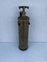 Front view of the antique brass fire extinguisher showing cylindrical shape, T-shaped top handle, and metal clasps.