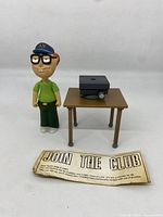 Family Guy figure standing next to a small wooden table holding a black projector and a paper card on surface