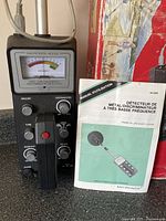 Front view of the Micronta 4000 metal detector unit with analog meter, control knobs, wired headphones, and user manual.