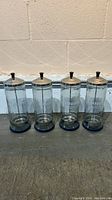 Four clear glass Barbicide jars with metal lids and black plastic stands on a concrete floor in front of a white wall. All jars are displayed upright.