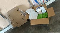 View of open cardboard box containing lime green crinkle paper, rainbow confetti tissue paper, and some packing supplies nearby, plus shrink-wrapped pallet of boxes in background.