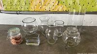 Seven assorted clear glass vases of various shapes and sizes displayed on a flat surface in front of a green and yellow background.