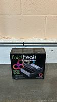 Photo of the Fold Freak Foil-it Dispenser in its original packaging, showing the box on a concrete floor against a beige wall.