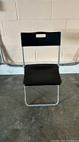 Photo of black folding chair showing full front view against a beige wall and concrete ground.