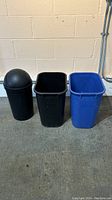 Three bins in a row against a wall: black bin with swinging lid, open black bin, blue recycling bin