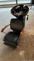 Front view of black hair washing basin with padded black chair and wooden armrests, showing basin and headrest attachment.