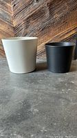 Photo showing two large round pots: a taller cream-colored one and a shorter black one, both with smooth matte finishes, placed side by side on a concrete surface with a wooden wall background.