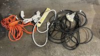 Five extension cords and power strips, varying colors including orange, white, and black, shown coiled and laid on a concrete floor.