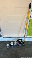 Photo shows a yellow-handled paint roller rod leaning against a wall next to five paint cans with different labels.