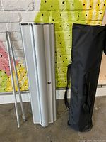 Photo showing disassembled banner stand base, two support poles, and black carrying case standing on floor against painted wall.