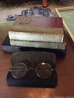Photo showing stack of three antique books with ornate cloth covers and a pair of vintage round glasses in an open black leather case.