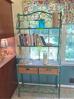 Front view of green metal bakers rack with wicker baskets and shelves with various kitchen and decor items.