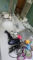Bathroom countertop with ceramic soap dispenser, tissue box cover, glass containers, and electrical appliances including hair dryer and irons with cords.