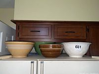 Four ceramic mixing bowls arranged in a row showing different colors and sizes: beige with stripes, green textured, brown striped, and white bowl with rim.