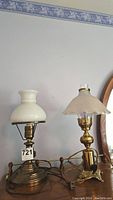 Both brass lamps shown side by side on a wooden surface below a plain wall, one with a smooth white glass shade and the other with a scalloped white glass shade.