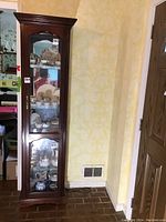 Full view of the wooden display cabinet standing against a yellow patterned wall, showing its tall and narrow profile with glass door and shelves inside.